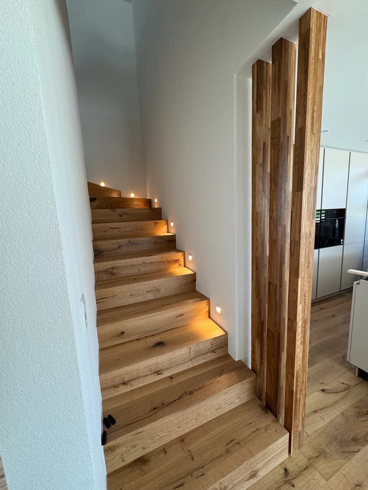 Beleuchtete Eichenholztreppe neben raumhohen Eichenlamellen und moderner Küche